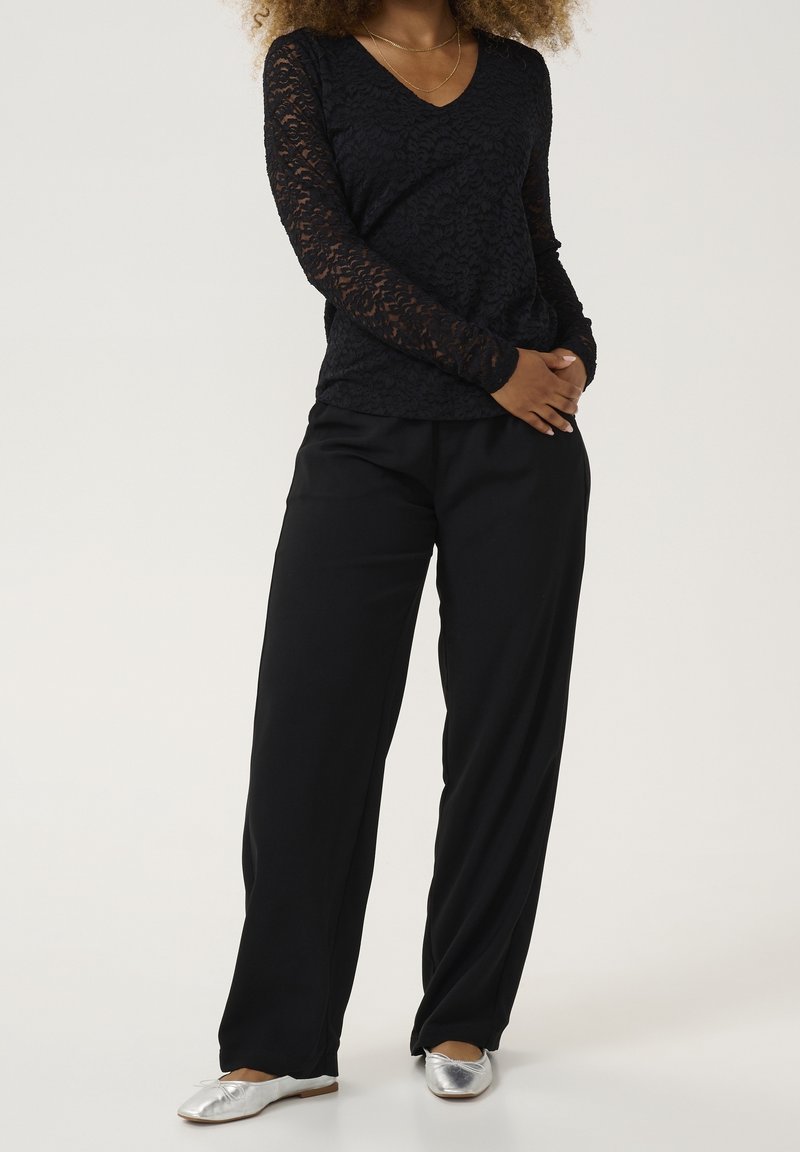 Top in pizzo nero a maniche lunghe con scollatura a V, abbinato a pantaloni neri a gamba larga e scarpe basse argentate. Dettagli in tessuto semplici e texturizzati.