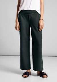 Kobieta ubrana w czarne spodnie o szerokich nogawkach, biały top bez rękawów, czarne klapki i srebrną bransoletkę na prawej ręce.