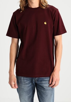 Mann trägt ein einfarbiges dunkelrotes T-Shirt mit einem kleinen gelben Logo auf der linken Brust und blaue Jeans, steht vor einem einfarbigen Hintergrund.