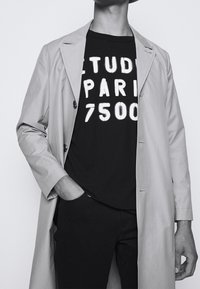Beige trench coat over a black graphic t-shirt displaying "ETUDES PARIS 7500" in white. Slim-fit black trousers. Minimalistic design.