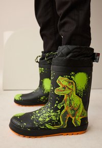 Stivali da pioggia in gomma nera con grafica di un dinosauro verde con dettagli a schizzi, polsino trapuntato e suola arancione con grip testurizzato.