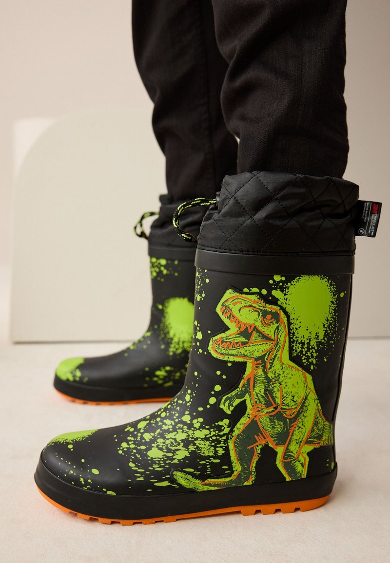 Bottes de pluie en caoutchouc noir avec un motif de dinosaure vert et des détails éclaboussés, une manchette matelassée et une semelle orange avec une prise texturée.