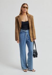 Hnědý semišový blejzr, černé tílko, světlé modré džíny s vysokým pasem a širokými nohavicemi, černá kabelka a tmavé sluneční brýle, stojící na jednobarevném pozadí.
