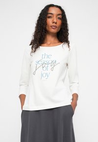 White long-sleeve shirt featuring blue and rhinestone text "the season of joy." Rounded neckline, smooth texture, worn with a grey skirt.