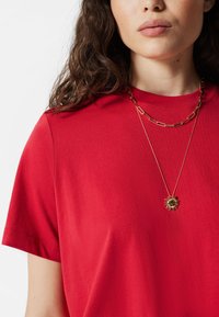 Rotes Baumwoll-T-Shirt mit kurzen Ärmeln, ausgestattet mit einer mehrlagigen goldenen Kettenkette und einem Sonnenblumen-Anhänger. Glatte Textur, einfaches Design.