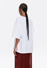 Camiseta blanca de gran tamaño hecha de algodón, con mangas cortas y un corte relajado. El modelo la lleva con una falda burgundy transparente.