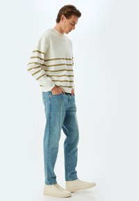 Sudadera blanca con rayas beige horizontales, combinada con vaqueros de un azul claro. El modelo lleva zapatillas beige, con las manos en los bolsillos, de perfil.