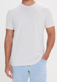 Lichtgrijs t-shirt met korte mouwen en een textuurpatroon; ronde halslijn; heeft een klein logo aan de linkerkant. Combinatie met lichtblauwe spijkerbroek.