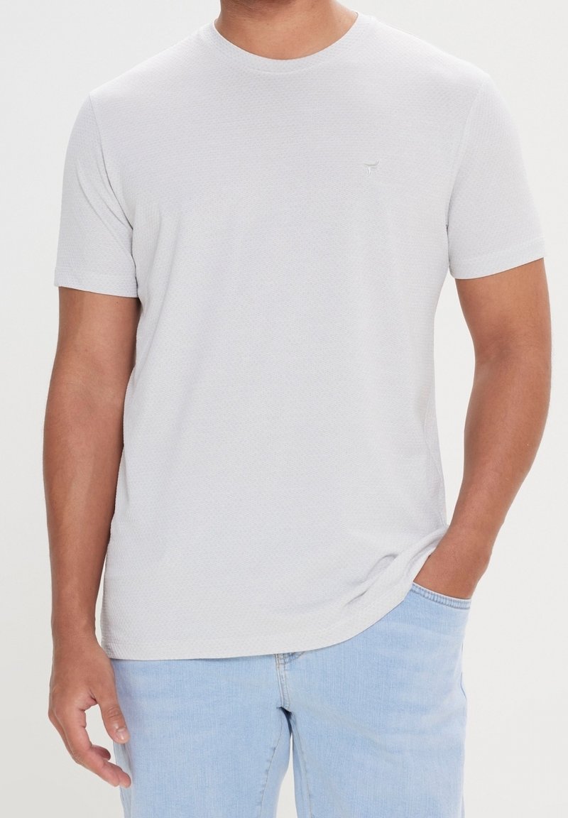 Lichtgrijs t-shirt met korte mouwen en een textuurpatroon; ronde halslijn; heeft een klein logo aan de linkerkant. Combinatie met lichtblauwe spijkerbroek.