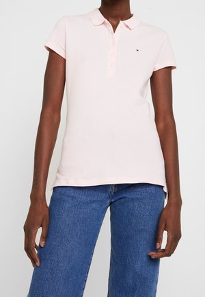 Person wearing a light pink short-sleeve polo shirt and blue denim jeans, standing against a plain white background.