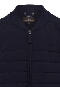 Marineblauwe gewatteerde bomberjack met een geribde kraag, volledige ritssluiting en geborduurd logo op de borst. Gladde stof met subtiele textuur.