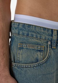 Světlé modré džíny z denimu s vybledlým efektem, oranžovým prošitím, malou černou značkou a bílým pasem, který je viditelný nad džíny.