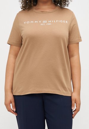 Osoba ubrana w beżowy t-shirt Tommy Hilfiger z białym logo i granatowe spodnie, stojąca na jednolitym tle.