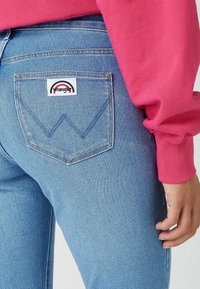 Světle modré džínové džíny s zadní kapsou, na které je bílá nášivka s červeným a modrým logem duhy. Kombinace s růžovým dlouhým rukávem.