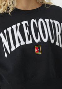 Personne aux cheveux bouclés portant un sweat-shirt noir avec un grand texte blanc "NIKECOURT" et un petit écusson du logo Nike multicolore en dessous.