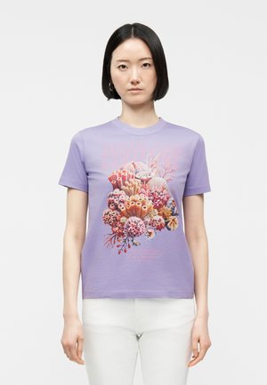 Mlada ženska s kratkimi črnimi lasmi, oblečena v lila majico z živobarvnim motivom koralnega grebena in bele hlače, stoji pred belim ozadjem.