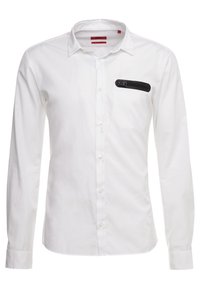 Witte blouse met lange mouwen en een button-down kraag, met een zwart ritszakje op de borst en klassieke knoopsluitingen.