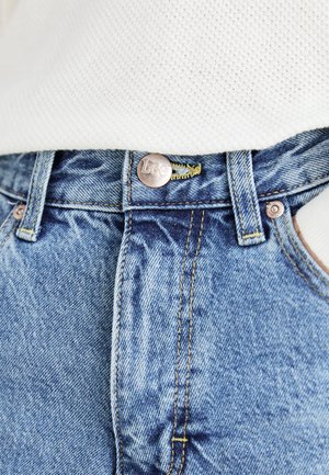 Nahaufnahme der Taille einer blauen Jeans aus Denim mit einem Metallknopf, auf dem "Lee" eingraviert ist, und gelben Nähten, kombiniert mit einem weißen, strukturierten Stoffoberteil.