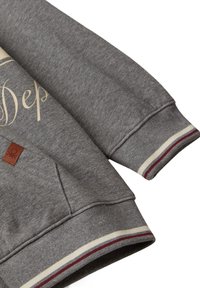 Grijze sweatshirtmouw en tailleband met crème en bordeauxrode strepen, gedeeltelijke beige schrifttekst en klein bruin leren merklabel.