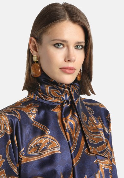 Satin bluza u mornarskoj plavoj boji s složenim zlatnim i smeškim uzorcima. Ima visoki kragnu i detalj sa vezivanjem, upotpunjena velikim jantar-naušnicama.