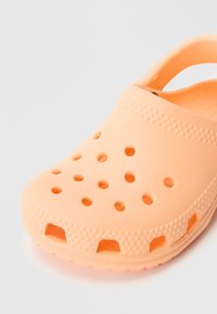 Persiku krāsas klogs ar ventilācijas caurumiem, strukturētu virsmu un aizmugurējo siksnu. Izgatavots no viegla materiāla, paredzēts komfortam un elpojamībai.