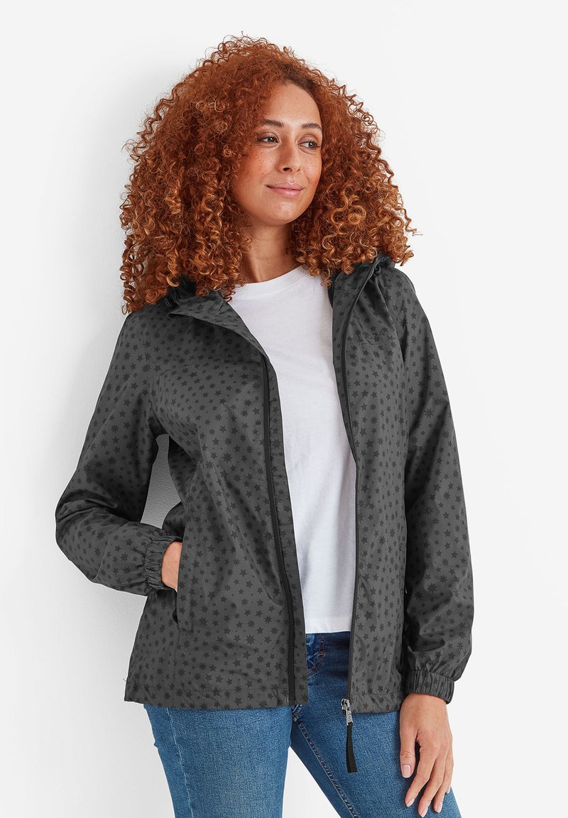 Woman with curly red hair wearing a grey star-patterned zip-up jacket, white shirt, and blue jeans, standing with hand in pocket.