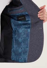 Ręka trzymająca otwartą ciemnoszary marynarkę, prezentującą niebieską podszewkę w wzór paisley oraz czarną metkę z napisem "BURATTI".