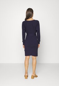 Femme aux longs cheveux bruns vêtue d'une robe bleu marine ajustée à manches longues et de sandales à talons carrés couleur camel, debout face à un mur blanc.