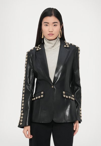 Schwarzer Lederblazer mit strukturiertem Design, goldenen Nietenakzenten entlang der Schultern und Ärmel sowie doppelten Fronttaschen. Weißer gestreifter Rollkragenpullover darunter.