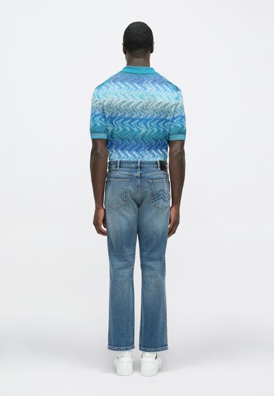 Rövid ujjú, kék mintás galléros póló hullám dizájnnal, világoskék farmerrel és fehér tornacipővel, hátulnézetből.