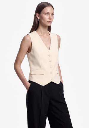 Donna che indossa un gilet beige senza maniche con bottoni e pantaloni neri a vita alta con le mani nelle tasche, guarda di lato su sfondo bianco.