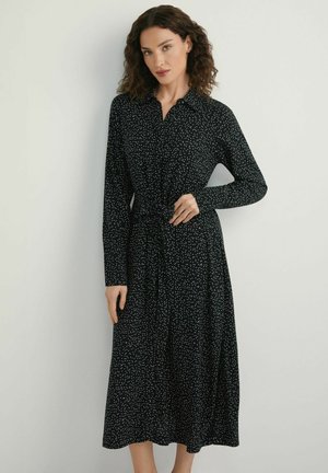 Kobieta o kręconych włosach, ubrana w czarną sukienkę z długim rękawem w białe kropki, z paskiem w talii, stojąca na tle jednolitego jasnego tła.