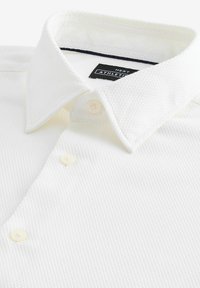 Weißes strukturiertes Poloshirt mit Kragen, das drei Knopfverschlüsse und ein mit Marineblau akzentuiertes Etikett im Halsbereich aufweist.