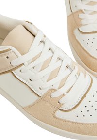 Sneakers bianchi e beige caratterizzati da un mix di pelle liscia e camoscio testurizzato, con lacci e perforazioni per la traspirabilità.