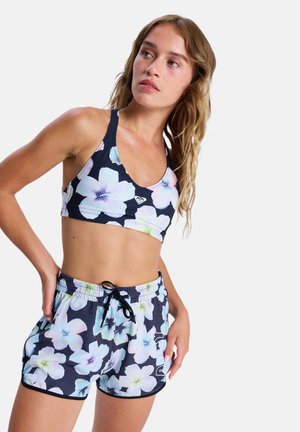 Młoda kobieta w dopasowanym czarnym staniku sportowym i krótkich spodenkach z dużym pastelowym wzorem kwiatowym, stojąca z rękami na biodrach na białym tle.