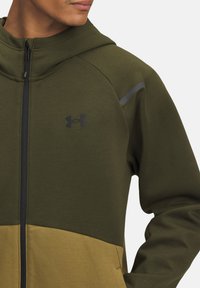 Haut à capuche zippé en vert foncé et olive, fabriqué en tissu doux. Il présente un panneau inférieur contrastant, des accents noirs et un logo Under Armour.