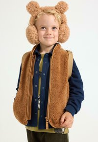Braune Fleece-Weste mit einem beigen Reißverschluss, getragen über einem blauen Hemd und einem grün gemusterten T-Shirt, kombiniert mit flauschigen beigen Ohrenwärmern.