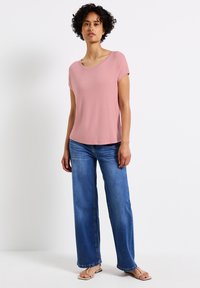 Růžový top s krátkým rukávem, volného střihu a širokým výstřihem, kombinovaný se světle modrými širokými džíny a béžovými sandály. Hladká textura materiálu.
