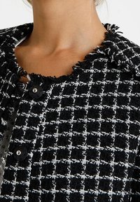 Czarna i biała teksturowana kurtka z wzorem w kratkę, strzępione krawędzie i okrągłe czarne guziki. Posiada klasyczny krój kołnierza.