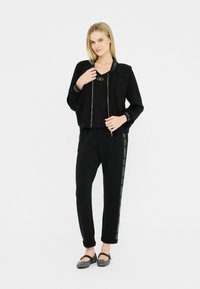 Ensemble de vêtements de sport noir comprenant une veste cropped zippée avec des accents de logo et un pantalon de jogging assorti avec une rayure logo sur le côté. Chaussures plates texturées.