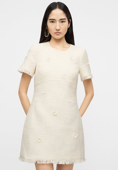 Νεαρή γυναίκα με ίσια σκούρα μαλλιά φορέσει κρεμ φόρεμα με κοντά μανίκια και υφή, με κρόσσια στα άκρα και μικρές ανθισμένες διακοσμήσεις.