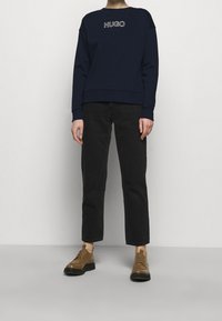 Persona che indossa una camicia a maniche lunghe blu navy con il testo "HUGO", pantaloni neri a gamba dritta e scarpe in pelle marrone con lacci, in piedi su uno sfondo semplice.