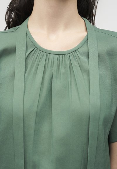 Κοντινό πλάνο ενός ατόμου που φοράει μια μπλούζα χρώματος φασκόμηλο με σούρες στη λαιμόκοψη και μακριές υφασμάτινες κορδέλες που κρέμονται μπροστά.