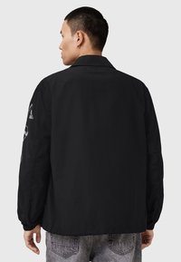 Giacca bomber leggera nera con maniche lunghe, polsini elasticizzati, colletto e dettagli intricati ricamati in bianco sulla manica sinistra.