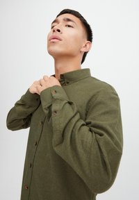 Cămașă verde măsliniu, cu nasturi, mâneci lungi, cu nasturi maro și guler. Fabricată dintr-un material moale, texturat.