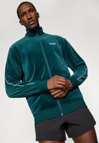 Tengeri zöld velúr tréningdzseki magas nyakú, elöl cipzárral és fehér oldalcsíkokkal. A mellkason PUMA logó található.