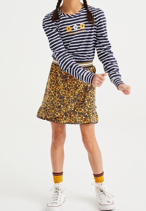 Meisje dat een gestreept shirt met lange mouwen met bloemenprint draagt, een gele bloemenrok, gele sokken en witte sneakers, staand met gebogen armen.