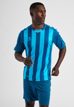 Mężczyzna biegnie w niebieskich szortach i sportowej koszulce w pionowe paski w jasnym i ciemnym odcieniu niebieskiego na jednolitym szarym tle.