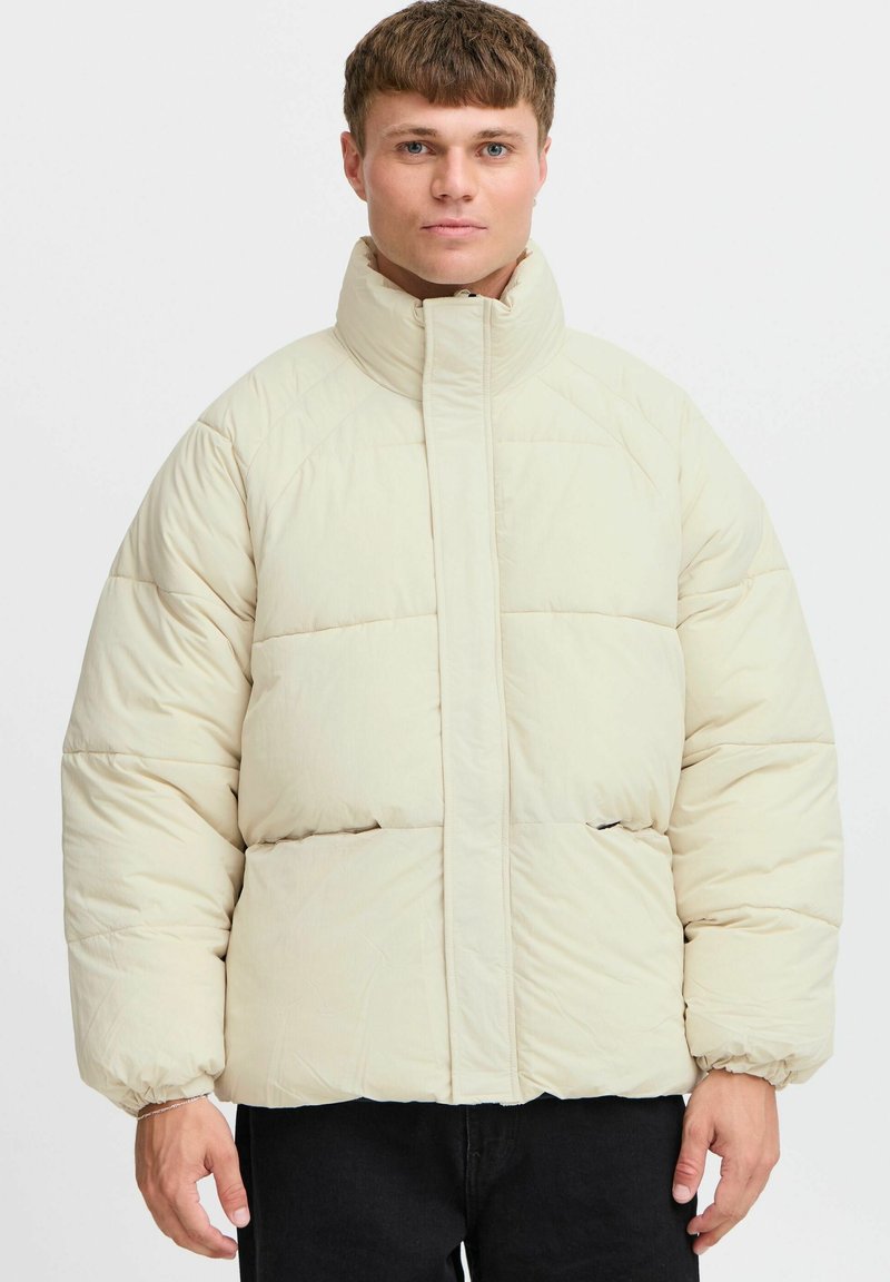 Beige pufferjakke med høj krave, quiltet design, elastiske manchetter og sidelommer, med en glat tekstur og afslappet pasform.