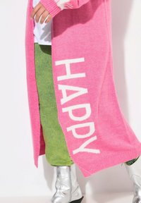 Cardigan rosa lavorato a maglia con la scritta "HAPPY" in bianco, abbinato a pantaloni verdi e stivaletti argentati, che mettono in risalto una texture morbida e colori vivaci.